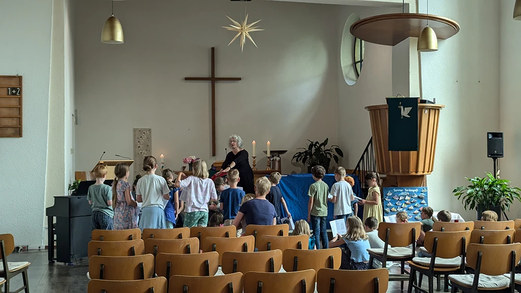 In der Kirche sind die Pfarrerin und viele aufmerksame Kinder, die gerade ein Lied proben.