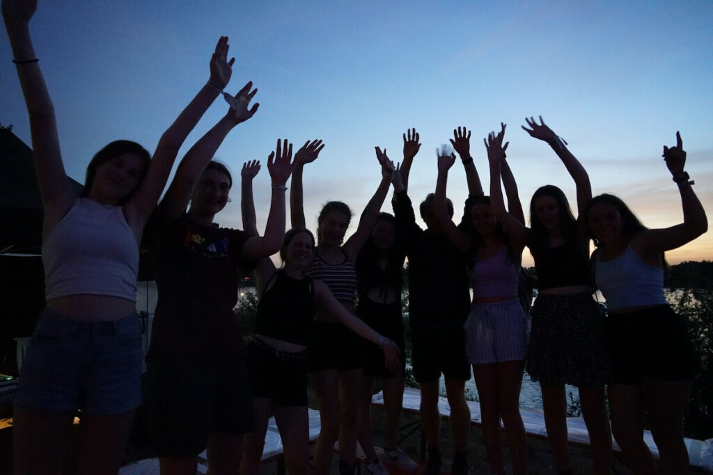 Man sieht eine Gruppe Jugendliche vor dem Abendhimmel, die Gesichter verschattet, die die Arme hoch reißen
