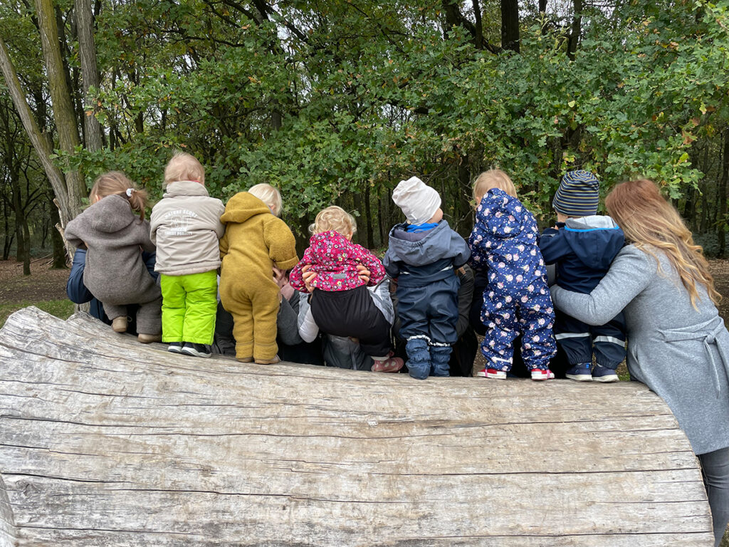 Viele Kinder stehen auf einem dicken Baumstamm. Sie werden zur Unterstützung fest gehalten, damit sie nicht hinfallen.