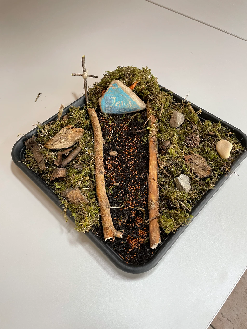 Minigarten in einer Schale mit Erde, Ästen, Moos, kleinen Steine und einem größeren Stein, auf dem "Jesus" steht.