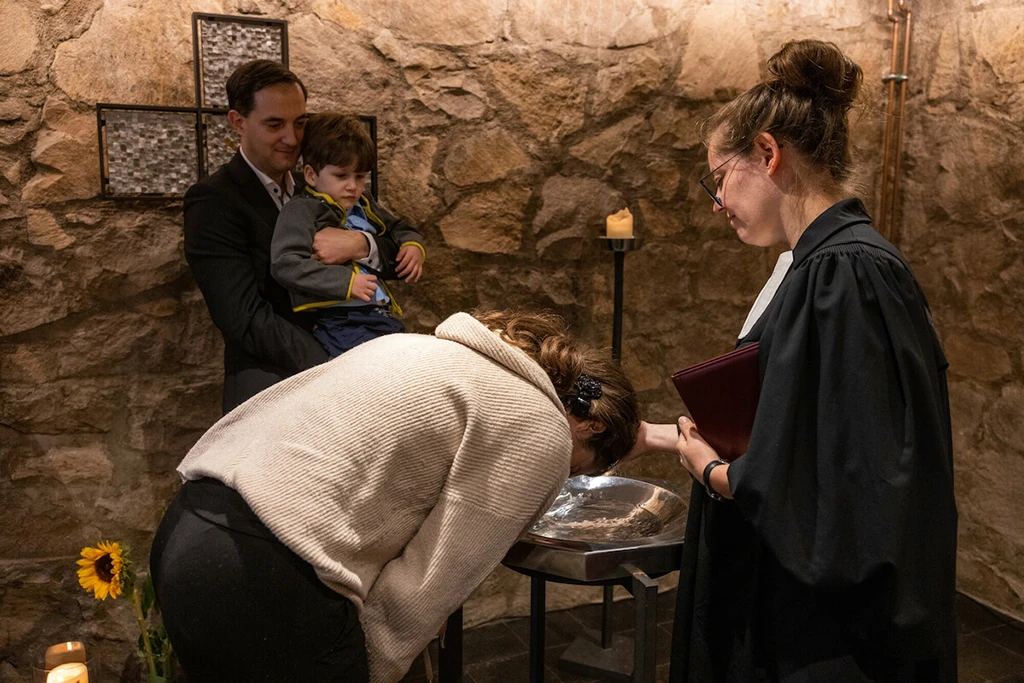 Eine Frau beugt sich für die Taufe über das Taufbecken. Ehemann und Sohn beobachten die Szene.